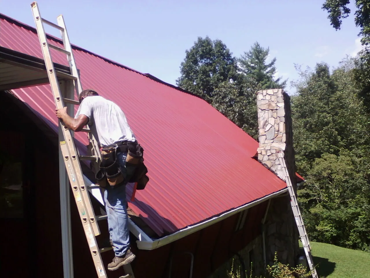 Roofer on a ladder beside a red metal roof and stone chimney — Soffit & Fascia Repair by Roofing Brothers in Brainard